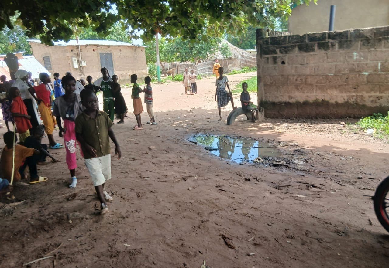 A rain-soaked earth path reflecting the sky; villagers walking between breeze-block houses.