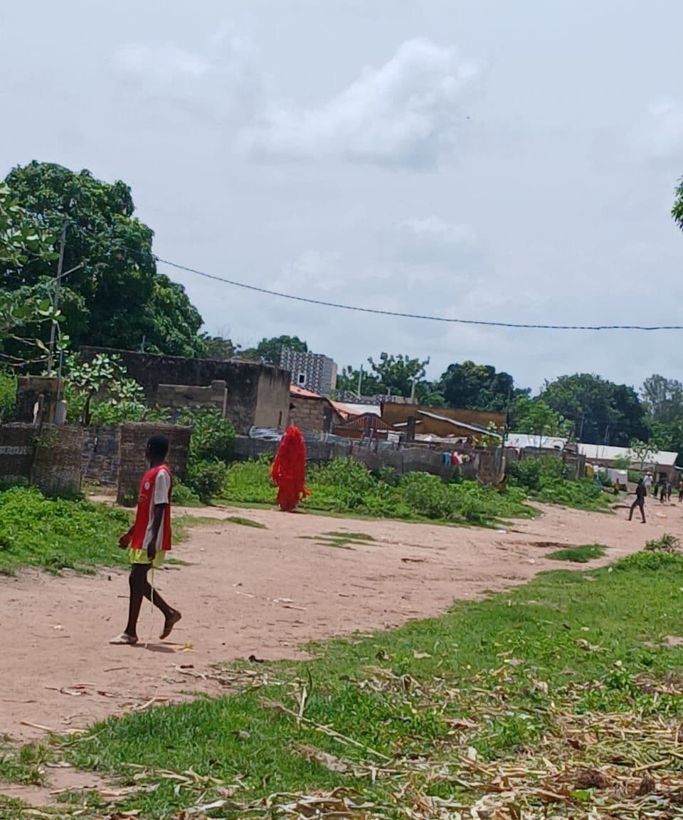A figure covered head to toe in red fibres walks along a sand track; a boy in a red jersey watches.